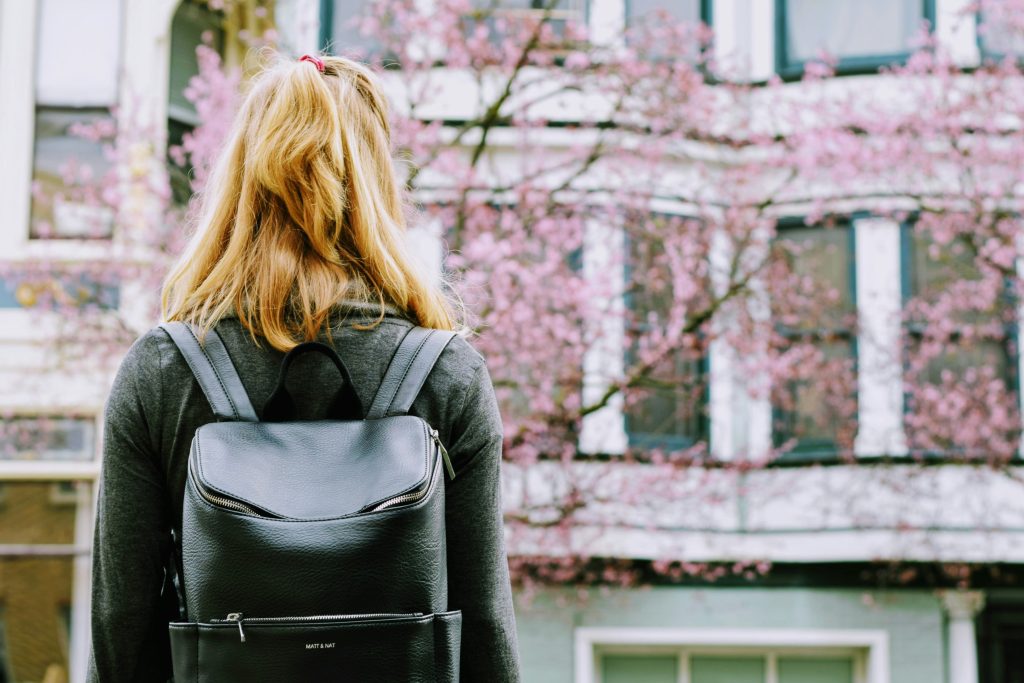 A person with shoulder length hair wearing a grey top and back backpack standing in from of pink cherry blossom tree