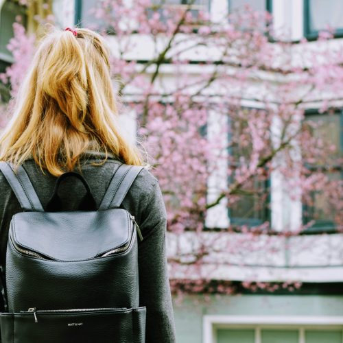 A person with shoulder length hair wearing a grey top and back backpack standing in from of pink cherry blossom tree A person with shoulder length hair wearing a grey top and back backpack standing in from of pink cherry blossom tree