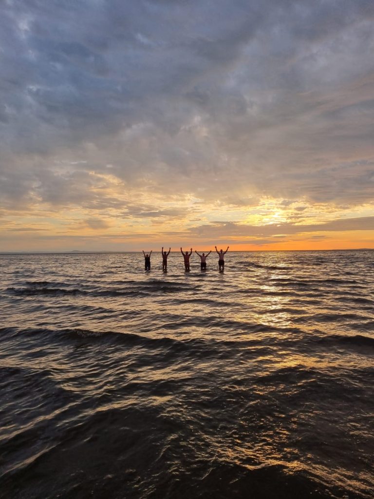 5 people standing in the distance in the water, hands up cheering, sunrise in the background